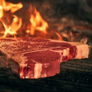 T-bone steak being grilled over open flames, showcasing sizzling outdoor barbecue cooking.